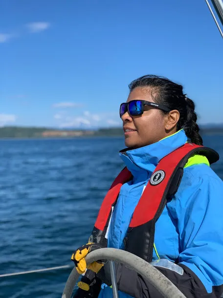 Urvashi in sunglasses and a red life jacket at the helm, looking out over the water